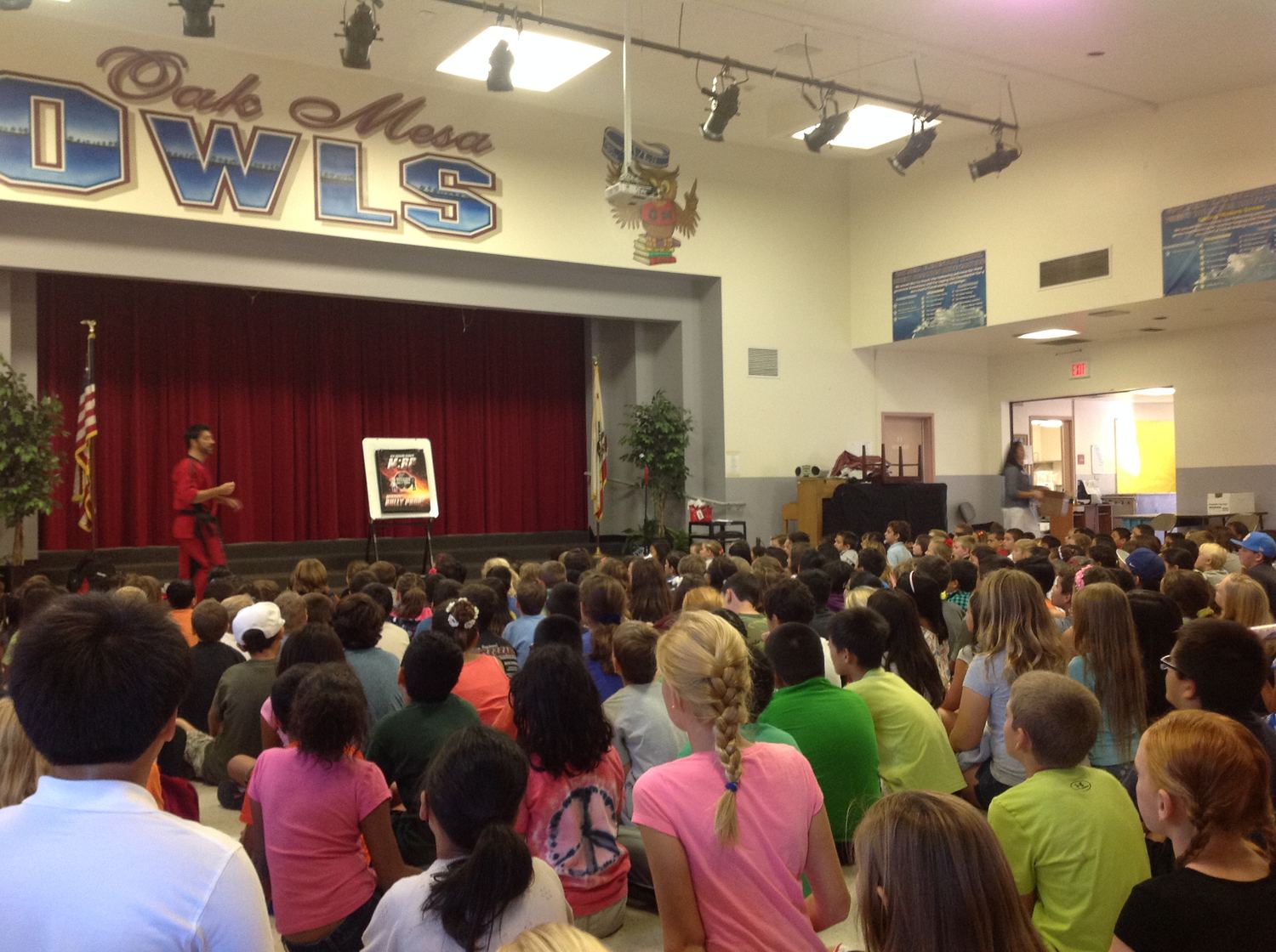 A school assembly hall filled with seated students facing a stage, where an instructor is presenting